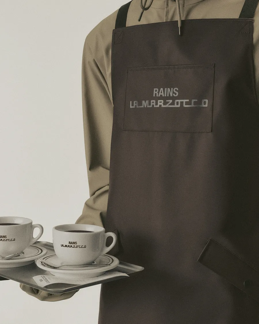 Brown Rains and La Marzocco barista apron with the logo on the chest pocket, holding a tray with two cups