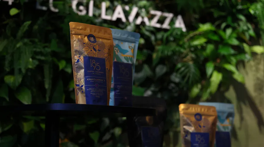 Coffee bags from 1895 Coffee Designers by Lavazza in front of a green plant wall with the Lavazza lettering